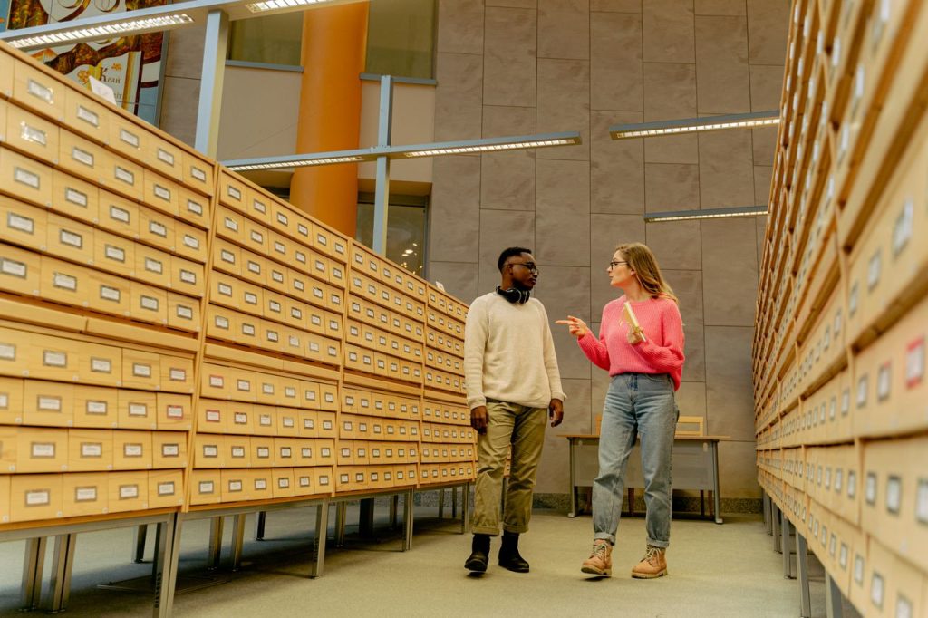Two adults conversing in a library archive aisle surrounded by wooden drawers and overhead lights.